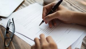 Unrecognizable businessman examining papers at table. Manager with ballpoint pen filling business papers. Close-up of male hands working at desk. Analyzing documents concept